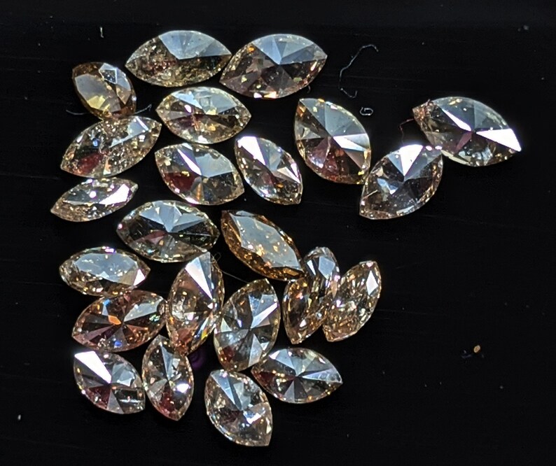 Puede incluir: Una colecci&oacute;n de diamantes de color marr&oacute;n claro de forma marquesa. Los diamantes est&aacute;n dispuestos en un mont&oacute;n suelto sobre una superficie negra.