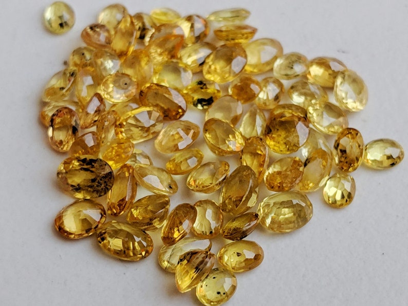 May include: A pile of oval-shaped, yellow gemstones. The gemstones are faceted and have a translucent appearance.