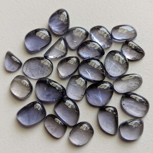 May include: A collection of 25 loose, faceted, purple-grey gemstones. The stones are smooth and have a slightly iridescent sheen.