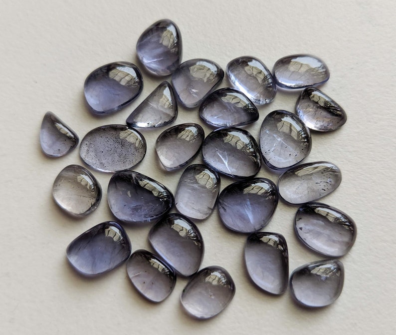 May include: A collection of 25 loose, faceted, purple-grey gemstones. The stones are smooth and have a slightly iridescent sheen.