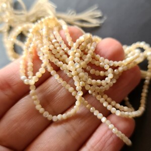 K&ouml;nnte beinhalten: Eine Str&auml;hne aus wei&szlig;en, irisierenden Muschelperlen, jede Perle ist facettiert und hat eine leicht unregelm&auml;&szlig;ige Form.