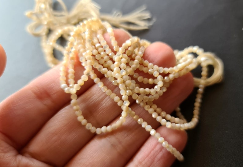 K&ouml;nnte beinhalten: Eine Str&auml;hne aus wei&szlig;en, irisierenden Muschelperlen, jede Perle ist facettiert und hat eine leicht unregelm&auml;&szlig;ige Form.