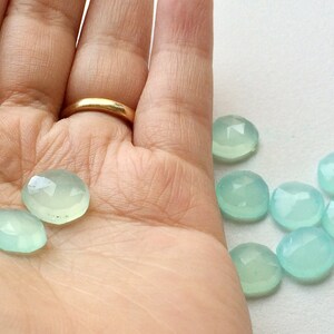 May include: A collection of faceted, pale blue-green gemstones. The gemstones are oval and round, with a polished, translucent appearance. Some are held in a hand, while others are scattered on a white surface. The gemstones have a subtle sparkle.