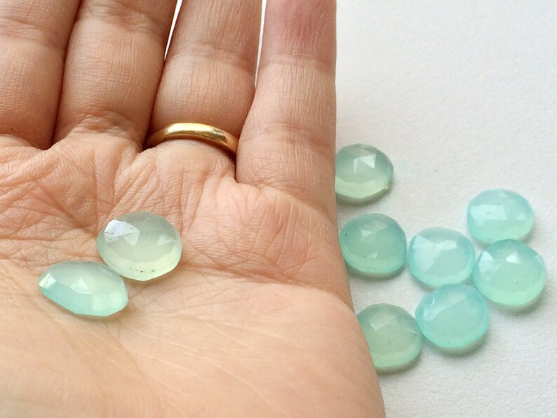 May include: A collection of faceted, pale blue-green gemstones. The gemstones are oval and round, with a polished, translucent appearance. Some are held in a hand, while others are scattered on a white surface. The gemstones have a subtle sparkle.