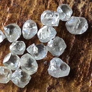 Könnte beinhalten: Eine Sammlung von rohen, ungeschliffenen Diamanten, auch bekannt als Rohdiamanten, die auf einer braunen Holzoberfläche präsentiert werden. Die Diamanten sind klar und haben verschiedene Formen und Größen.