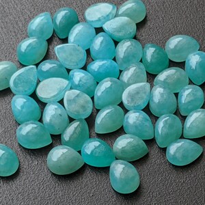 May include: A collection of numerous loose, teardrop-shaped gemstones in varying shades of translucent teal and light blue. Each stone features a smooth, polished, cabochon-style surface that reflects light, creating a glossy appearance across the group.