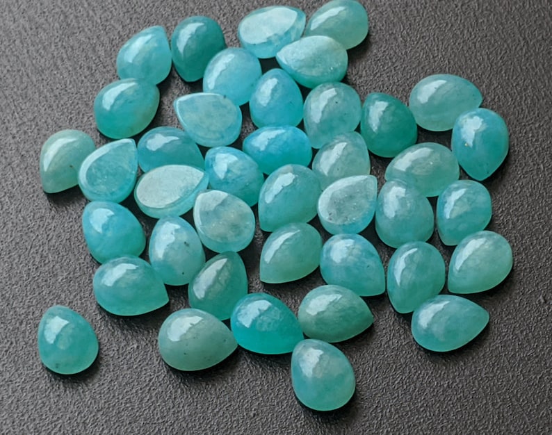 May include: A collection of numerous loose, teardrop-shaped gemstones in varying shades of translucent teal and light blue. Each stone features a smooth, polished, cabochon-style surface that reflects light, creating a glossy appearance across the group.