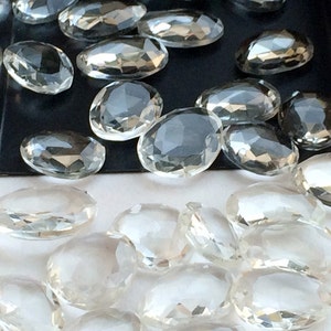Peut inclure: Une collection de cristaux de quartz transparents, facet&eacute;s et de forme ovale. Les cristaux sont dispers&eacute;s sur une surface blanche et un plateau noir.
