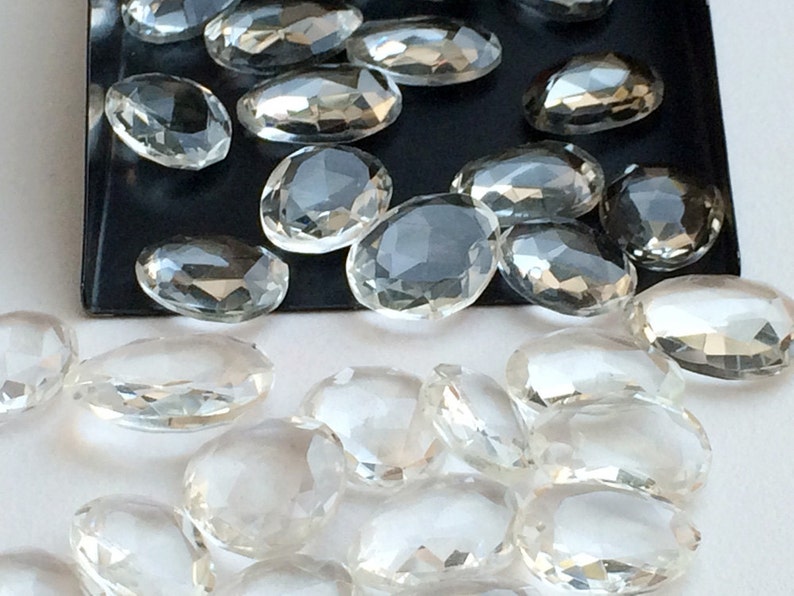 Peut inclure: Une collection de cristaux de quartz transparents, facet&eacute;s et de forme ovale. Les cristaux sont dispers&eacute;s sur une surface blanche et un plateau noir.