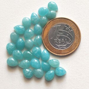 May include: A collection of small, teardrop-shaped, light blue gemstones arranged next to a one-real coin for scale. The stones have a smooth, polished surface and exhibit subtle variations in their opaque, cyan-toned coloration.