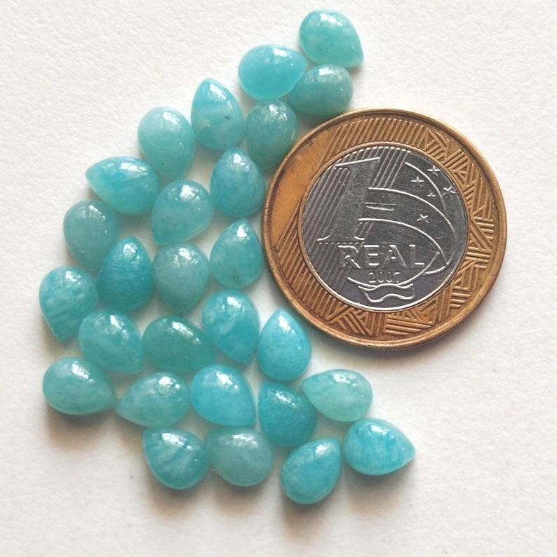 May include: A collection of small, teardrop-shaped, light blue gemstones arranged next to a one-real coin for scale. The stones have a smooth, polished surface and exhibit subtle variations in their opaque, cyan-toned coloration.