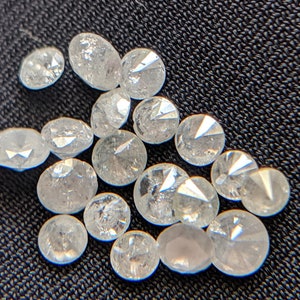 May include: A collection of small, clear, faceted, round, and oval-shaped diamonds. The diamonds are scattered on a dark gray fabric surface.