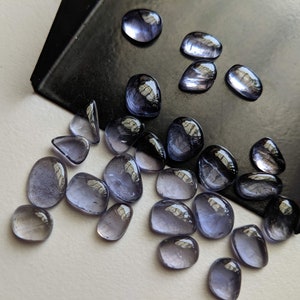 May include: A collection of 20 small, smooth, oval-shaped, iridescent, purple-blue gemstones. The gemstones are scattered on a white surface.