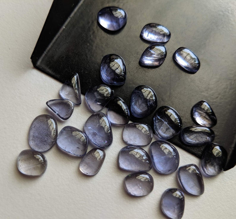 May include: A collection of 20 small, smooth, oval-shaped, iridescent, purple-blue gemstones. The gemstones are scattered on a white surface.