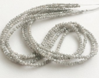 May include: A strand of gray diamond beads. The beads are small and have a rough, faceted texture. The strand is long and is draped over a white surface.