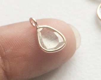 May include: A small, clear teardrop-shaped pendant with a rose gold setting. The pendant is on a finger.