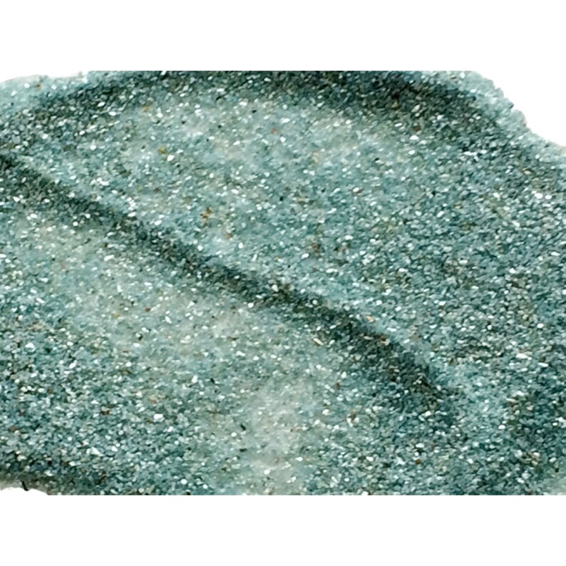 May include: A close-up of teal blue glitter with a shimmery finish. The glitter is spread out in a loose, uneven layer, creating a textured effect.