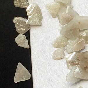 May include: A collection of small, clear, iridescent, faceted crystals. The crystals are scattered on a white surface.