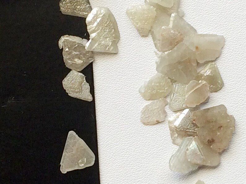 May include: A collection of small, clear, iridescent, faceted crystals. The crystals are scattered on a white surface.