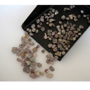 May include: A close-up of a pile of small, rough, pink and brown diamonds. The diamonds are scattered on a white surface and in a black tray.