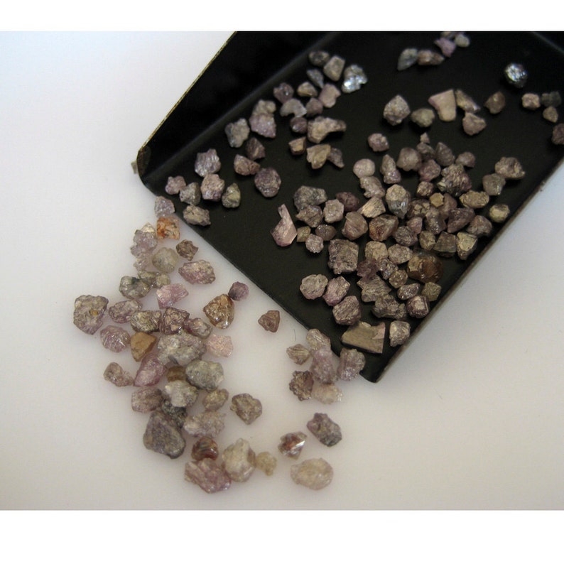 May include: A close-up of a pile of small, rough, pink and brown diamonds. The diamonds are scattered on a white surface and in a black tray.