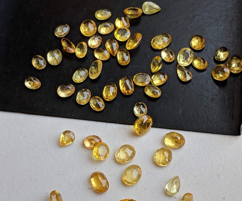 May include: A collection of yellow gemstones, likely citrine or topaz, in various oval and pear shapes. The stones are scattered on a black surface.