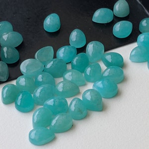 May include: A collection of numerous small, smooth, pear-shaped turquoise gemstones with a polished finish. The stones are scattered across a surface, with some resting inside a black metal scoop, displaying a consistent vibrant blue-green color.