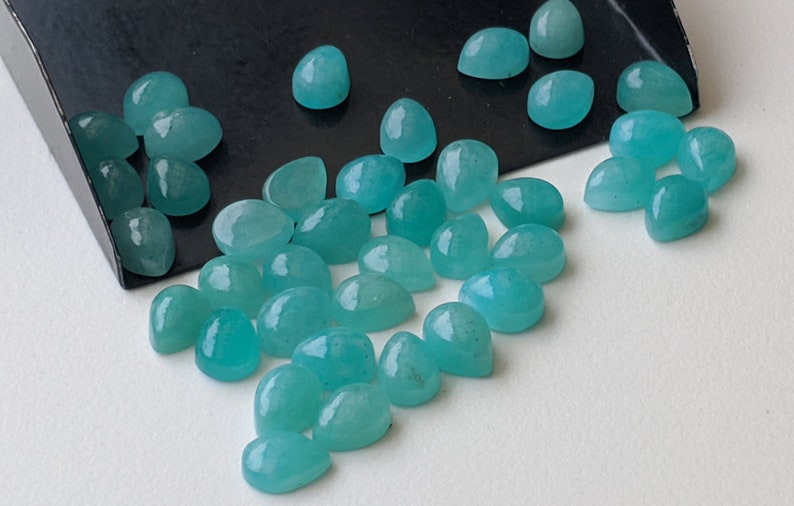 May include: A collection of numerous small, smooth, pear-shaped turquoise gemstones with a polished finish. The stones are scattered across a surface, with some resting inside a black metal scoop, displaying a consistent vibrant blue-green color.