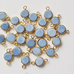 Può includere: Una collezione di 20 perline rotonde di pietra preziosa di colore blu chiaro con connettori a doppio anello dorati. Le perline sono sparse su una superficie bianca.