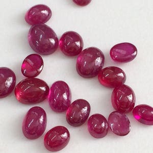 May include: A collection of oval-shaped, polished ruby gemstones in various sizes. The rubies are a deep red color and have a glossy finish.