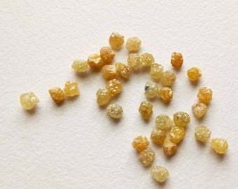 Puede incluir: Una colección de pequeños diamantes amarillos en bruto. Los diamantes están dispersos sobre una superficie blanca.