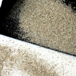 May include: A black scoop with a pile of fine, light brown sand on top. The sand is spilling out of the scoop onto a white surface.