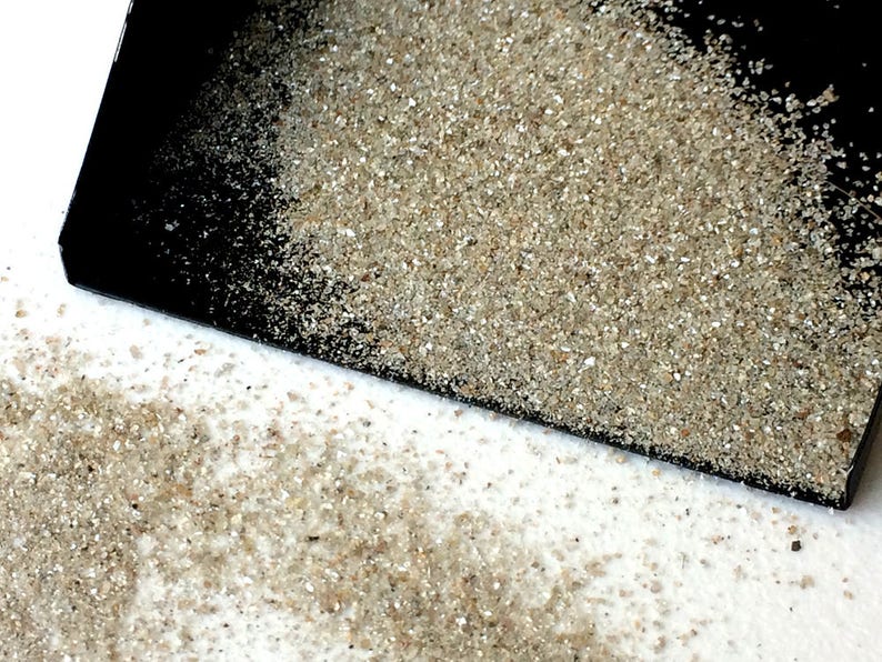 May include: A black scoop with a pile of fine, light brown sand on top. The sand is spilling out of the scoop onto a white surface.