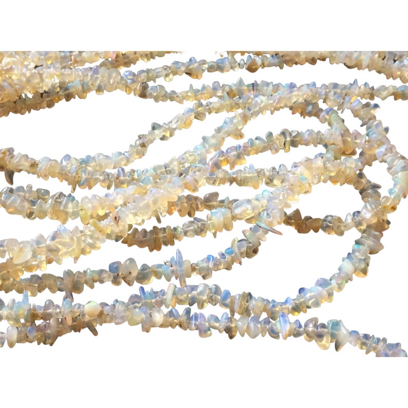 May include: A strand of iridescent white opal chip beads. The beads are small and irregular in shape, and they are strung on a clear thread. The beads are scattered and overlapping, creating a textured and organic look.