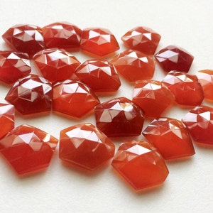 Puede incluir: Una colecci&oacute;n de 20 piedras de cornalina facetadas de color rojo-naranja. Las piedras est&aacute;n dispuestas en un mont&oacute;n suelto sobre una superficie blanca.