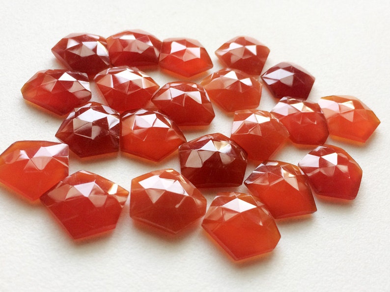 Puede incluir: Una colecci&oacute;n de 20 piedras de cornalina facetadas de color rojo-naranja. Las piedras est&aacute;n dispuestas en un mont&oacute;n suelto sobre una superficie blanca.