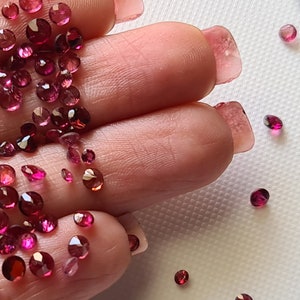 Op de afbeelding: Een close-up van een hand die een grote hoeveelheid losse roze en rode edelstenen vasthoudt. De edelstenen zijn van verschillende groottes en vormen, en sommige zijn verspreid over een wit oppervlak.