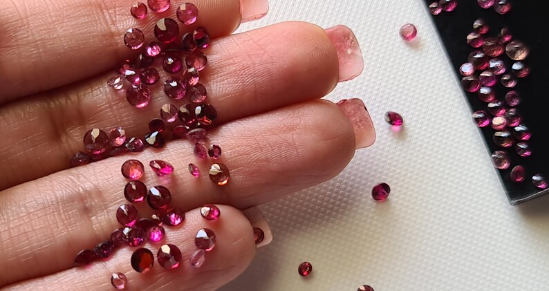 Op de afbeelding: Een close-up van een hand die een grote hoeveelheid losse roze en rode edelstenen vasthoudt. De edelstenen zijn van verschillende groottes en vormen, en sommige zijn verspreid over een wit oppervlak.