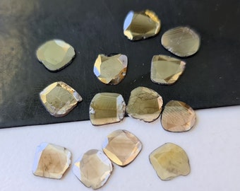 May include: A collection of faceted, irregularly shaped gemstones. The stones have a golden-brown hue with reflective surfaces. The shapes vary from rounded to more angular, with visible facets. The gemstones are scattered on a light surface.