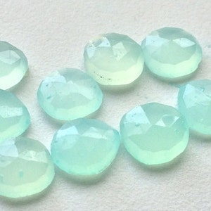 May include: A collection of nine faceted, light blue-green gemstones. Each stone has a rounded shape with multiple facets, creating a sparkling effect. The gems are arranged on a white surface.