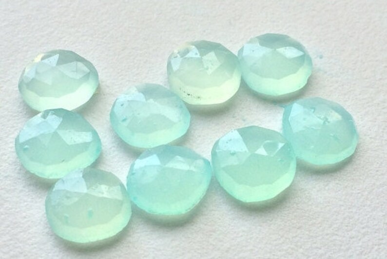 May include: A collection of nine faceted, light blue-green gemstones. Each stone has a rounded shape with multiple facets, creating a sparkling effect. The gems are arranged on a white surface.