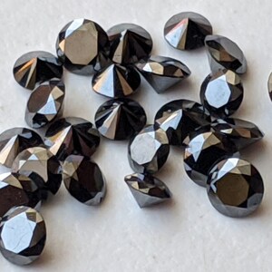 May include: A collection of black round diamonds, also known as carbonado diamonds, with a faceted cut. The diamonds are scattered on a white surface.