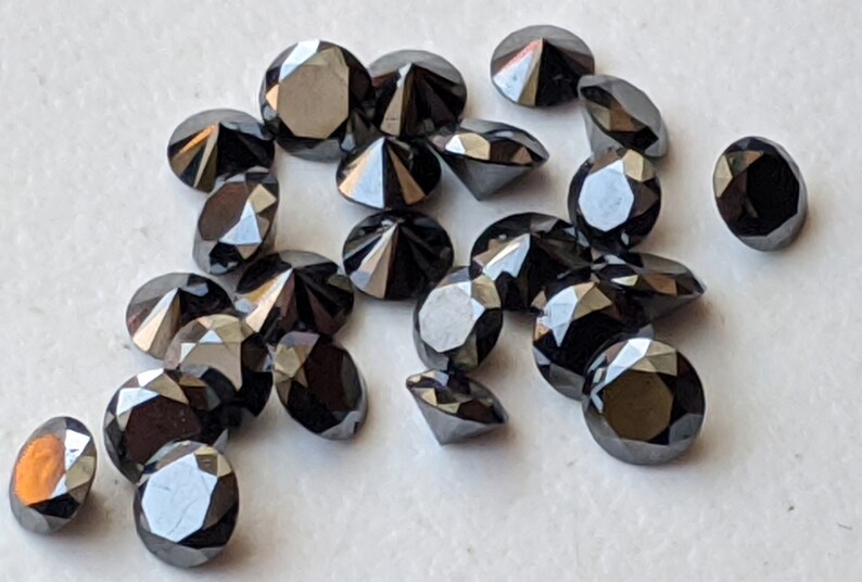 May include: A collection of black round diamonds, also known as carbonado diamonds, with a faceted cut. The diamonds are scattered on a white surface.