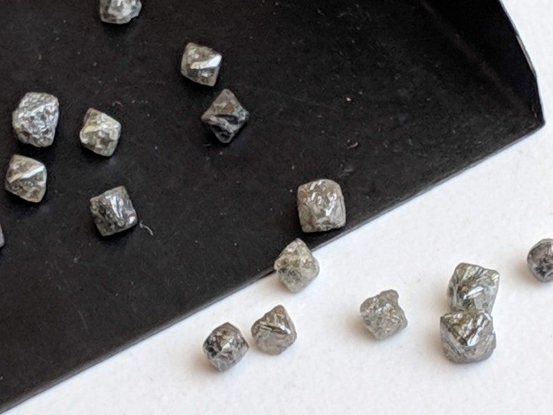Puede incluir: Una colecci&oacute;n de peque&ntilde;as piedras grises, &aacute;speras, con forma de diamante sobre una superficie negra. Las piedras est&aacute;n dispersas sobre una superficie blanca.