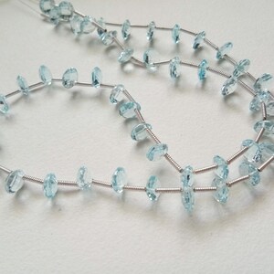 May include: A strand of light blue gemstone beads strung on silver wire. The beads are oval shaped and have a faceted finish.