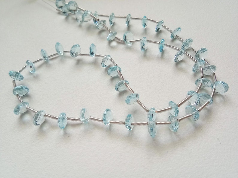 May include: A strand of light blue gemstone beads strung on silver wire. The beads are oval shaped and have a faceted finish.