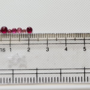 Op de afbeelding: Een rij van acht ronde glazen kralen in roze en rood van verschillende groottes, gemeten met een liniaal in centimeters.