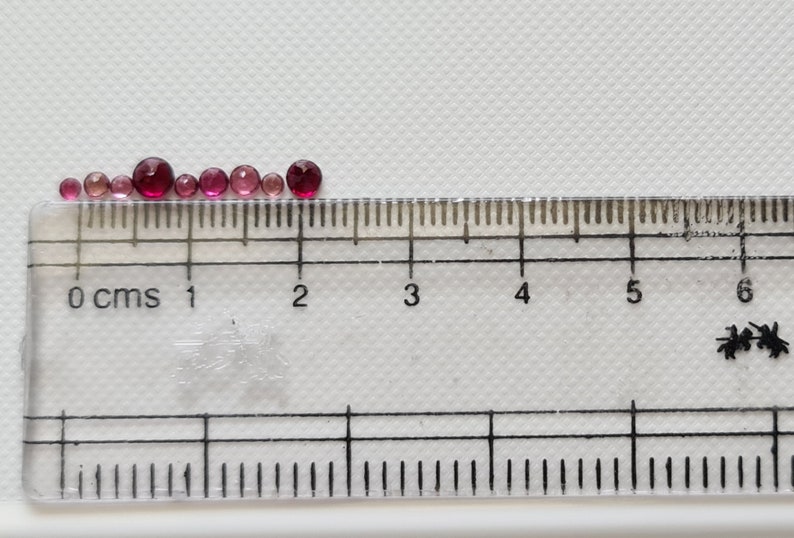 Op de afbeelding: Een rij van acht ronde glazen kralen in roze en rood van verschillende groottes, gemeten met een liniaal in centimeters.