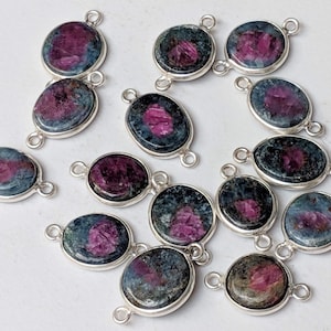 May include: A collection of 17 oval-shaped gemstone pendants with silver jump rings. The gemstones are a mix of green, blue, and pink.