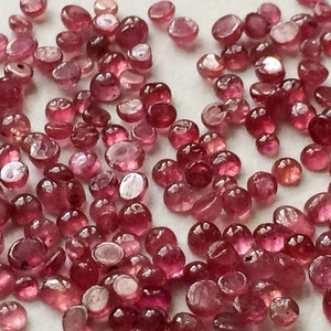 Puede incluir: Un gran grupo de piedras preciosas redondas y facetadas de color rosa. Las piedras preciosas están esparcidas sobre una superficie blanca.